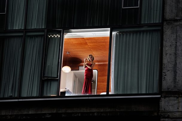 woman in long red dress holding a glass of wine and looking down of a window