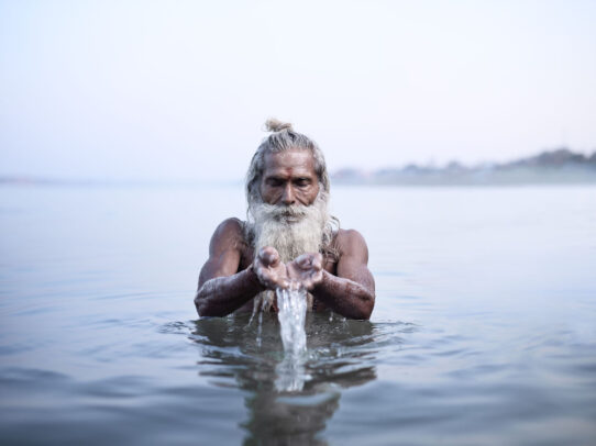 old man with white beard picking up water from the river in India