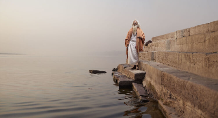 old man with a stick on steps in front of a river in India