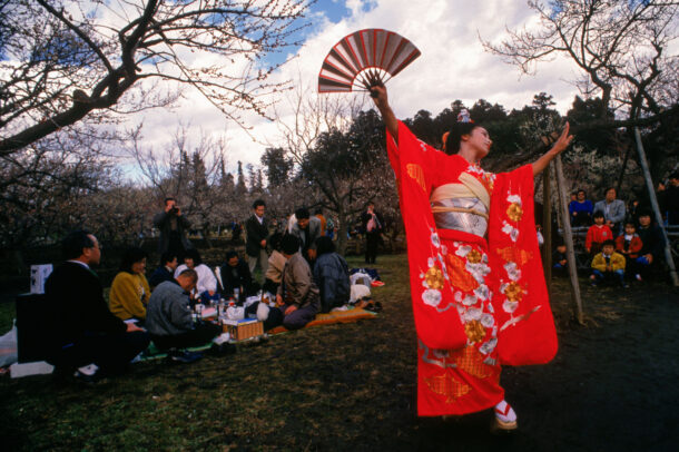 japanese woman in kimono holding a fan dancing in a park with people 